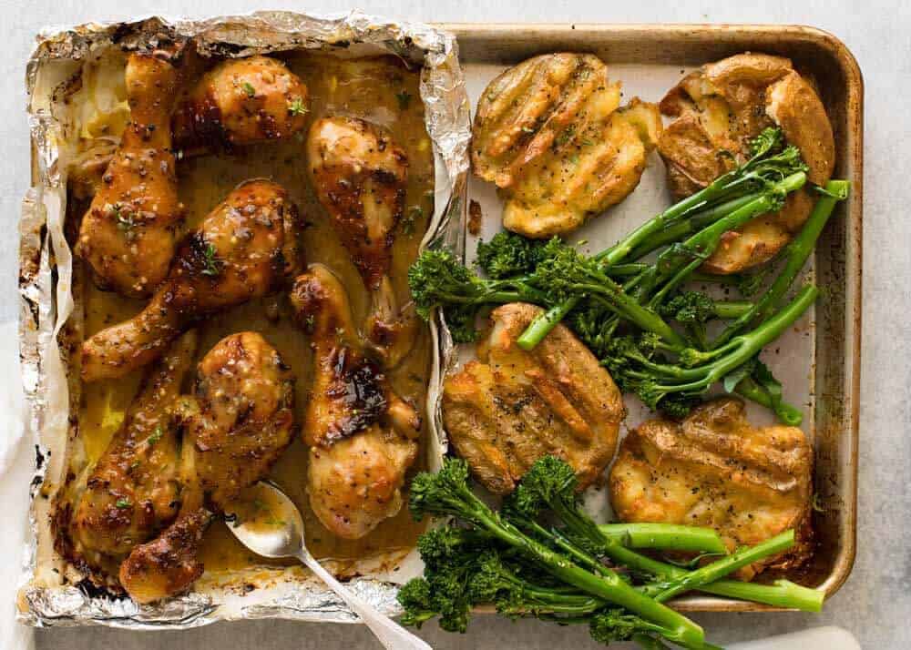 Overhead photo of Honey Mustard Baked Chicken Drumsticks with Smashed Potatoes on a silver tray with a side of steamed broccolini