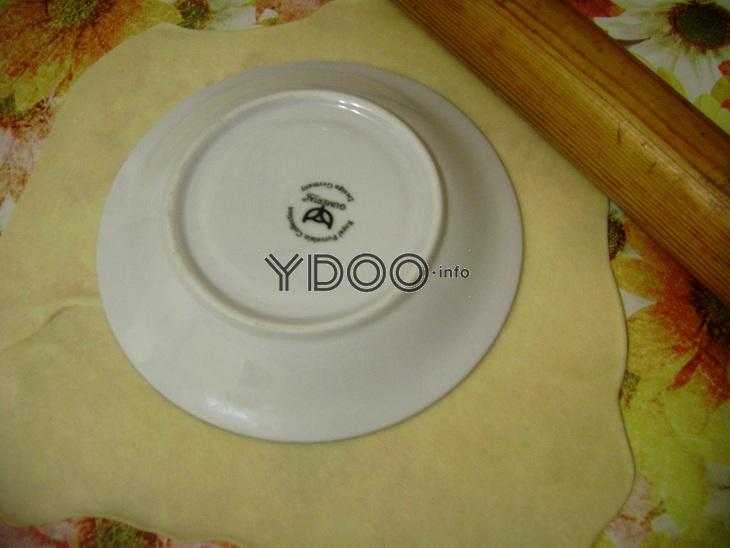 на раскатанном пласте теста лежит круглая тарелка, рядом деревянная скалка